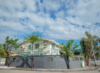 這張圖片展示一幢位於低密度住宅區的兩層洋房，建築物外圍設有灰色圍牆，旁邊有街道路口、路牌、樹木及街燈。畫面天氣晴朗，天空開揚，整體呈現新界低密度屋苑的寧靜居住氛圍，適合用於介紹 錦綉花園、洋房生活、家庭置業及低密度住宅環境 的文章封面。