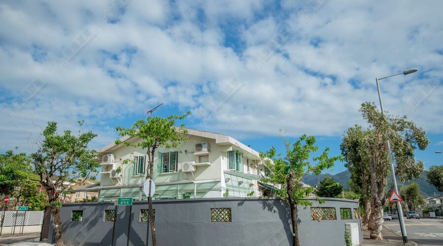 這張圖片展示一幢位於低密度住宅區的兩層洋房，建築物外圍設有灰色圍牆，旁邊有街道路口、路牌、樹木及街燈。畫面天氣晴朗，天空開揚，整體呈現新界低密度屋苑的寧靜居住氛圍，適合用於介紹 錦綉花園、洋房生活、家庭置業及低密度住宅環境 的文章封面。